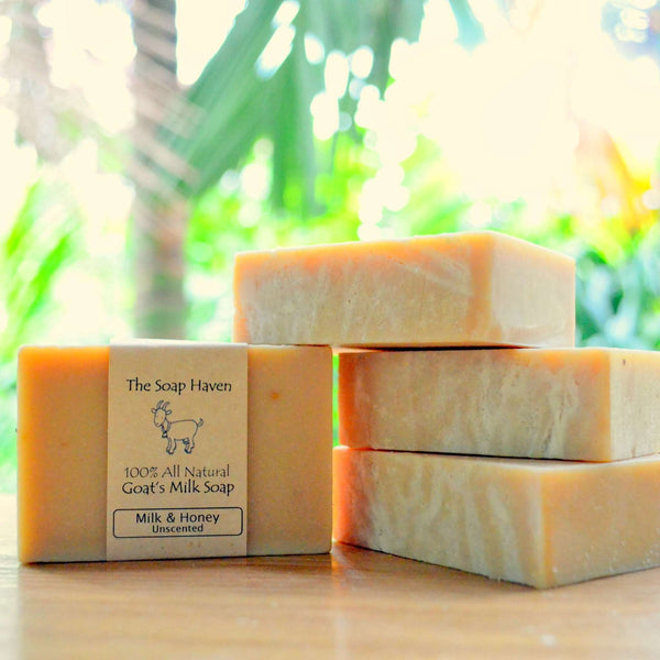 Four bars of goat milk soap stacked on a wooden surface, with one bar wrapped in a paper label that reads 'The Soap Haven 100% All Natural Goat's Milk Soap Milk & Honey Unscented.'